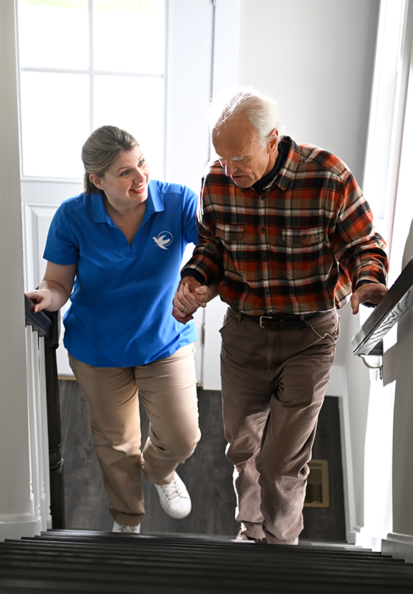 A Visiting Angels caregiver guiding an elderly man as he uses a walker, showcasing fall prevention measures in a supportive home setting.